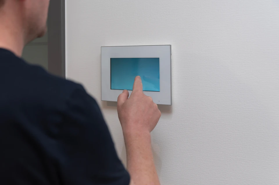 Homme en veste noire pointant du doigt un écran tactile mural affichant un plan d'étage 2D d'une maison